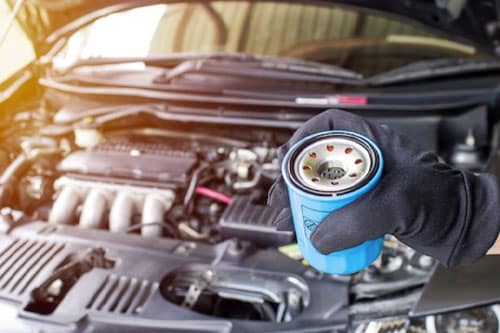 A gloved hand holds an oil filter in front of an open car hood with the engine visible.