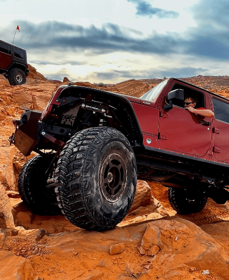 A red off-road vehicle from the Barney Brothers dealership in Grand Junction conquers a rocky terrain, while the driver leans out the window; another vehicle is visible in the background.