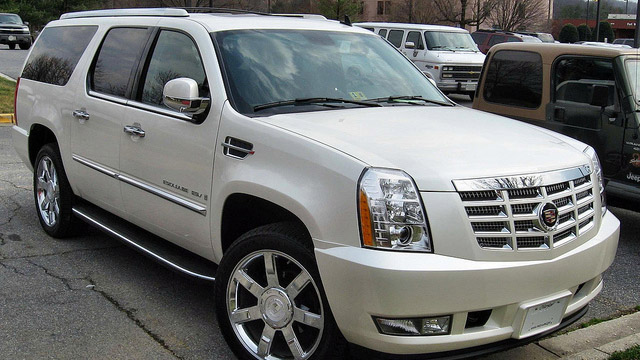 A Cadillac Escalade SUV, striking in its white finish, is parked on a street with a building and other vehicles in the background.