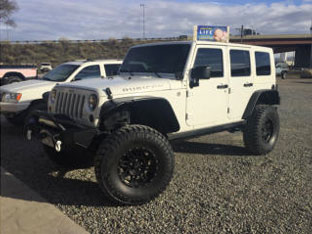 A white Jeep Wrangler with oversized tires, one of the impressive customer rigs, parked on a gravel surface, with another vehicle in the background.
