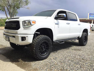 White lifted pickup truck parked on gravel with large off-road tires and black rims. Trees and a signboard in the background showcase one of our impressive customer rigs.