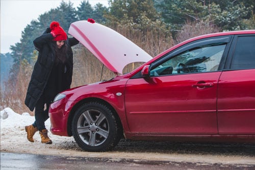 A person in winter clothing peers under the open hood of a red car on a snowy roadside, likely checking the battery.