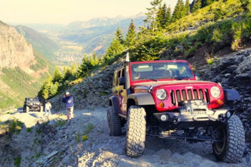 Red off-road vehicle on a rocky mountain trail with people walking nearby; scenic view of mountains and valley behind. Keep an eye out—black bears are known to wander near the road.