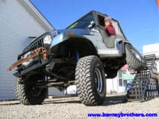 A blue off-road vehicle with large tires is on a ramp; a person is inside, showcasing one of our impressive customer rigs. Visit www.barneybrothers.com for more details.