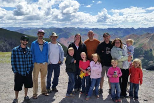 A group of 14 people, including kids, stands on a mountain overlook with a scenic backdrop of clouds and peaks during their company trip.