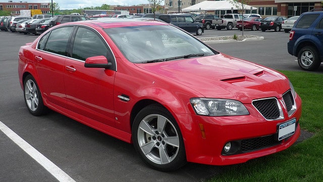 A classic red Pontiac sedan is parked in a lot among other cars, with a building standing proudly in the background.