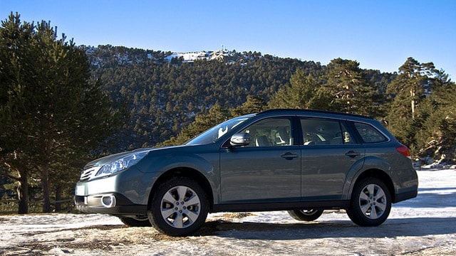 A blue Subaru station wagon is parked on a snowy landscape, with majestic mountains and trees in the background.