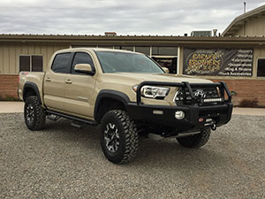 A beige, lifted Toyota truck is parked on gravel in front of a matching beige building under a cloudy sky.