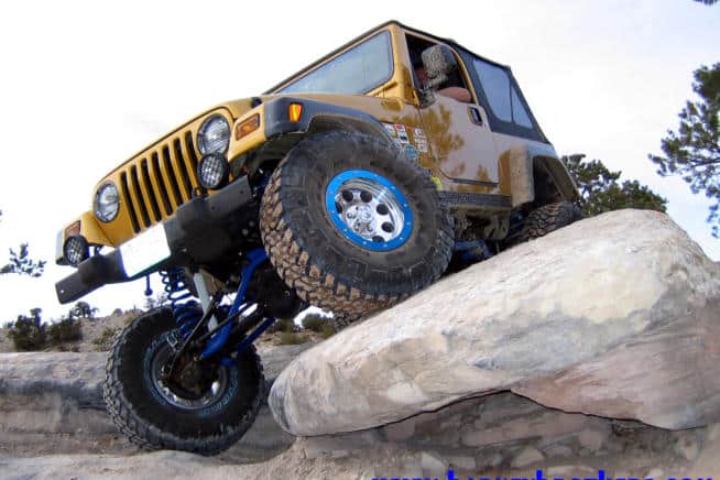 A yellow off-road vehicle, equipped with rugged off-road wheels, climbs over large rocks in a rocky outdoor landscape.