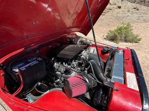 The hood of a red car is open, revealing the engine and various components inside, seemingly poised for some engine repair. A grassy landscape is in the background.