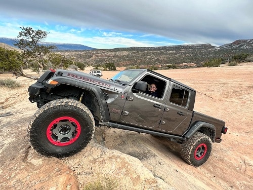 A grey Jeep Rubicon with striking red off-road wheels climbs a rocky terrain; a person is visible through the open front window, set against a rugged desert backdrop.