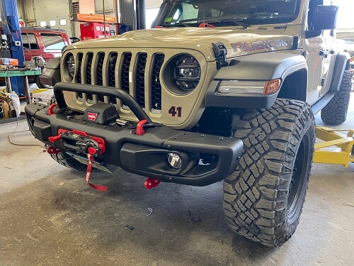 A rugged Jeep Rubicon with off-road tires, off-road accessories, and a front winch is parked in a garage for maintenance.