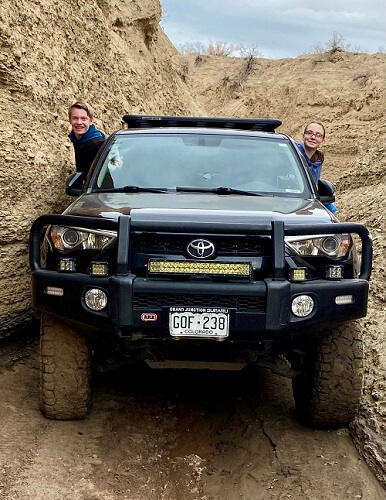 Two people smiling from the windows of a Toyota SUV with off-road mods, navigating a narrow, rocky canyon trail.