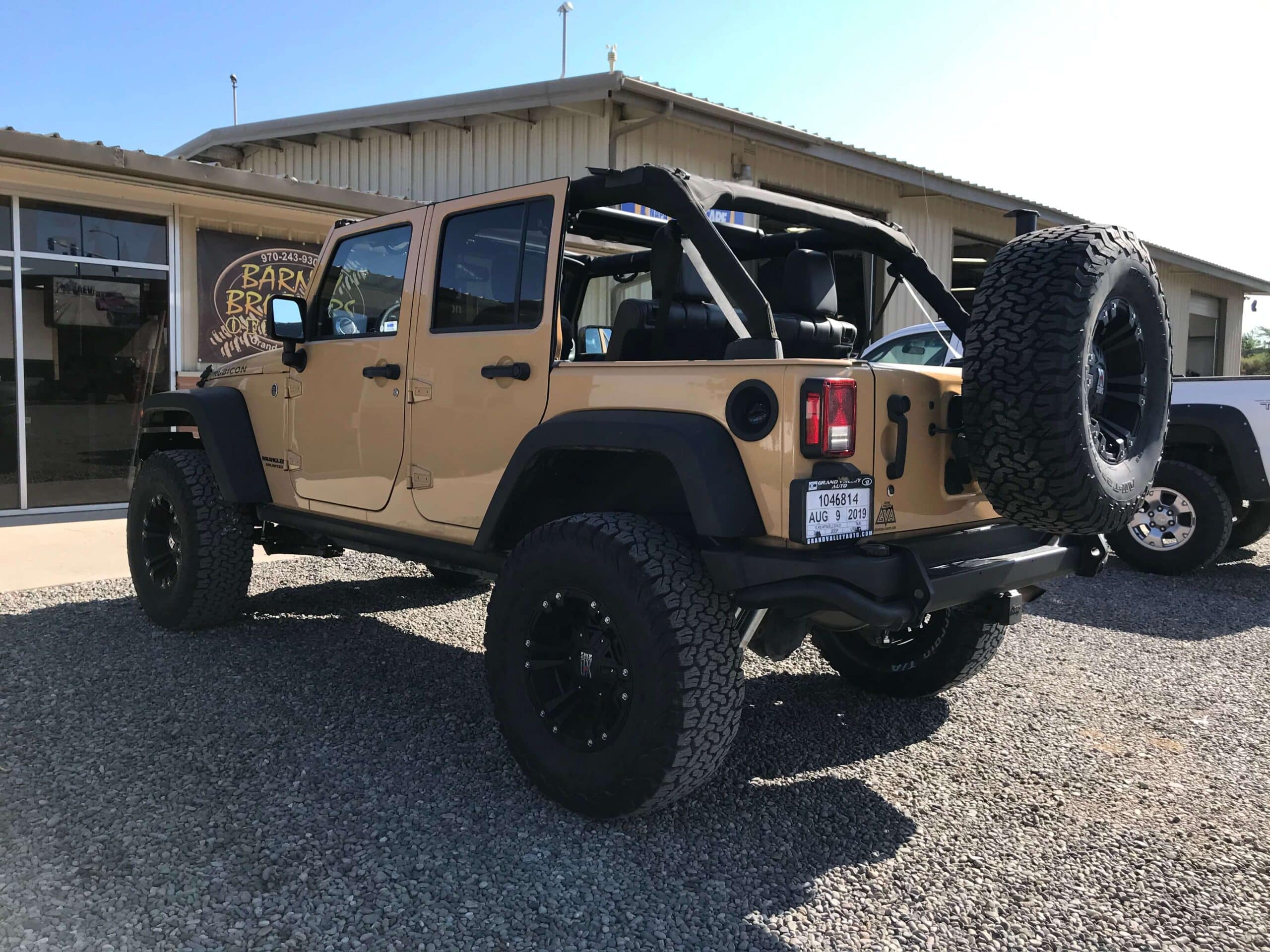 A tan, four-door Jeep Wrangler with off-road mod tires is parked outside a building with a sign that reads "Barn.