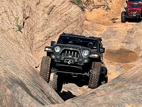 A black off-road vehicle, equipped with rugged off-road accessories, navigates a rocky, narrow terrain with a red vehicle trailing in the background.