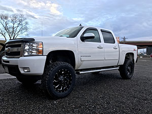 A lifted white Chevrolet Silverado pickup truck with black wheels is parked on a gravel lot under a cloudy sky, embodying rugged elegance and dependable performance.