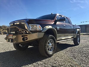 A lifted, maroon Dodge pickup truck with large tires is parked on a gravel surface under a clear sky.