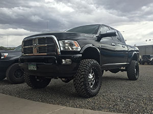 A black Dodge pickup truck with large off-road tires is parked on a gravel surface under a cloudy sky.
